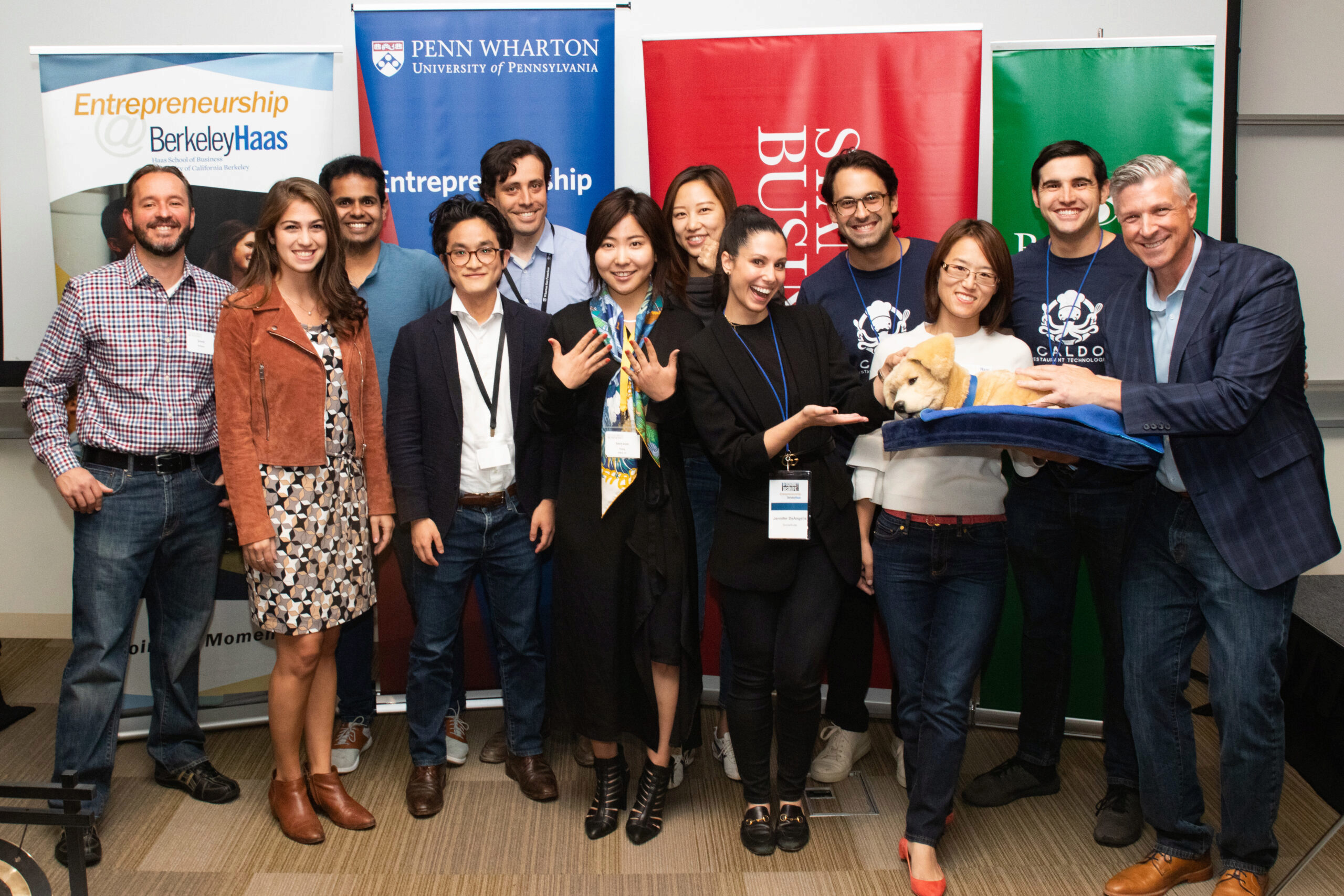 A group of people stand smiling in front of banners from various business schools. One person holds a small dog on a blue blanket.