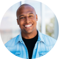 Headshot of a person smiling, wearing a denim jacket and black shirt, with a blurred outdoor background.